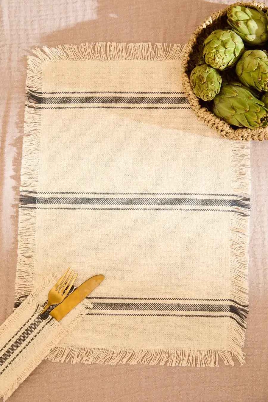 Set de table en coton épais beige et avec rayure gris foncé tissée et finition frangée. Des artichauts posés dessus et des couverts.