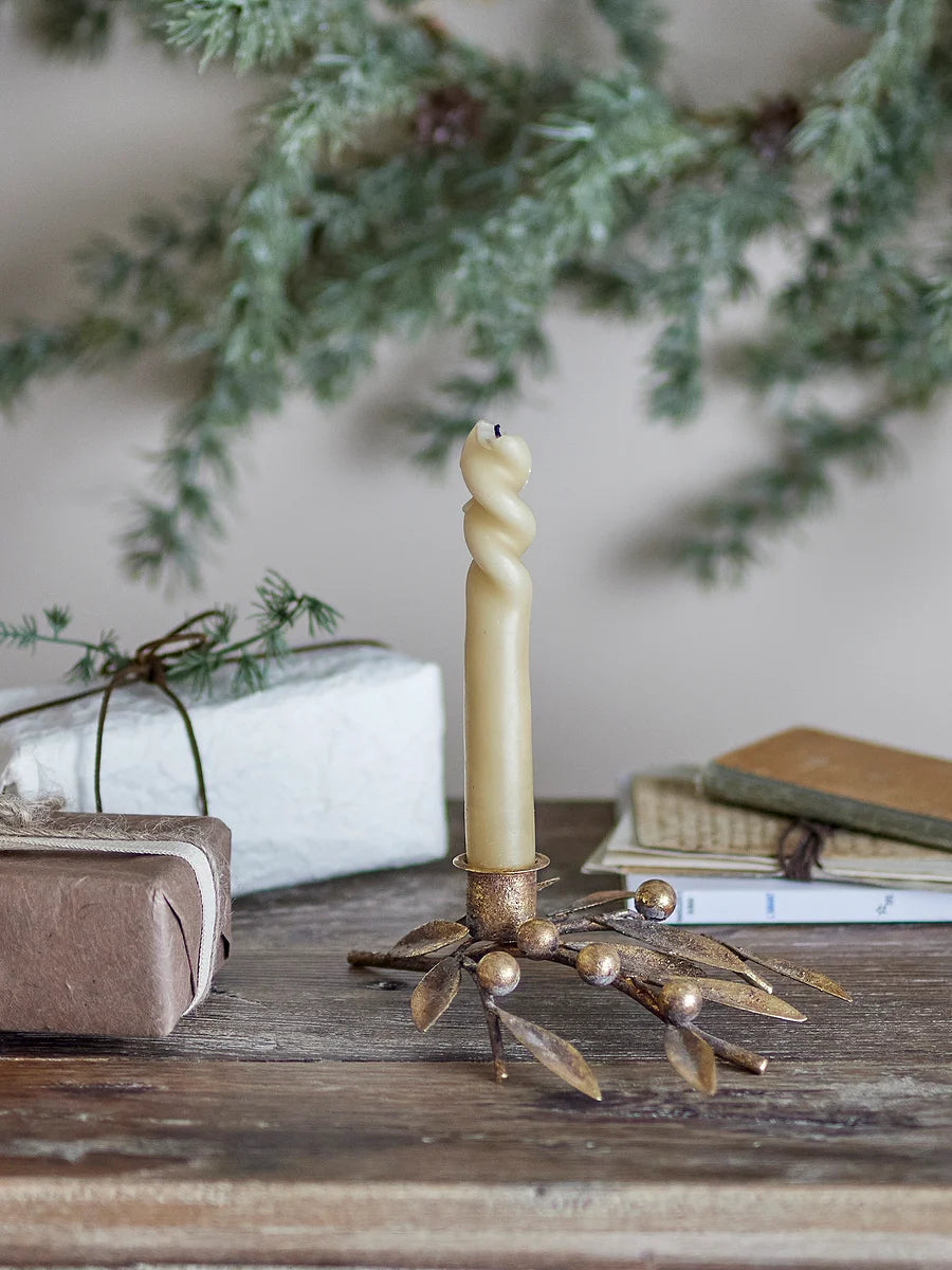 Bougeoir en métal finition or vieilli décoré de branches de gui, pour bougies longues de 2,2 cm de diamètre. avec une bougie beige torsadée. Posé sur une table en bois dans un décor de Nöel