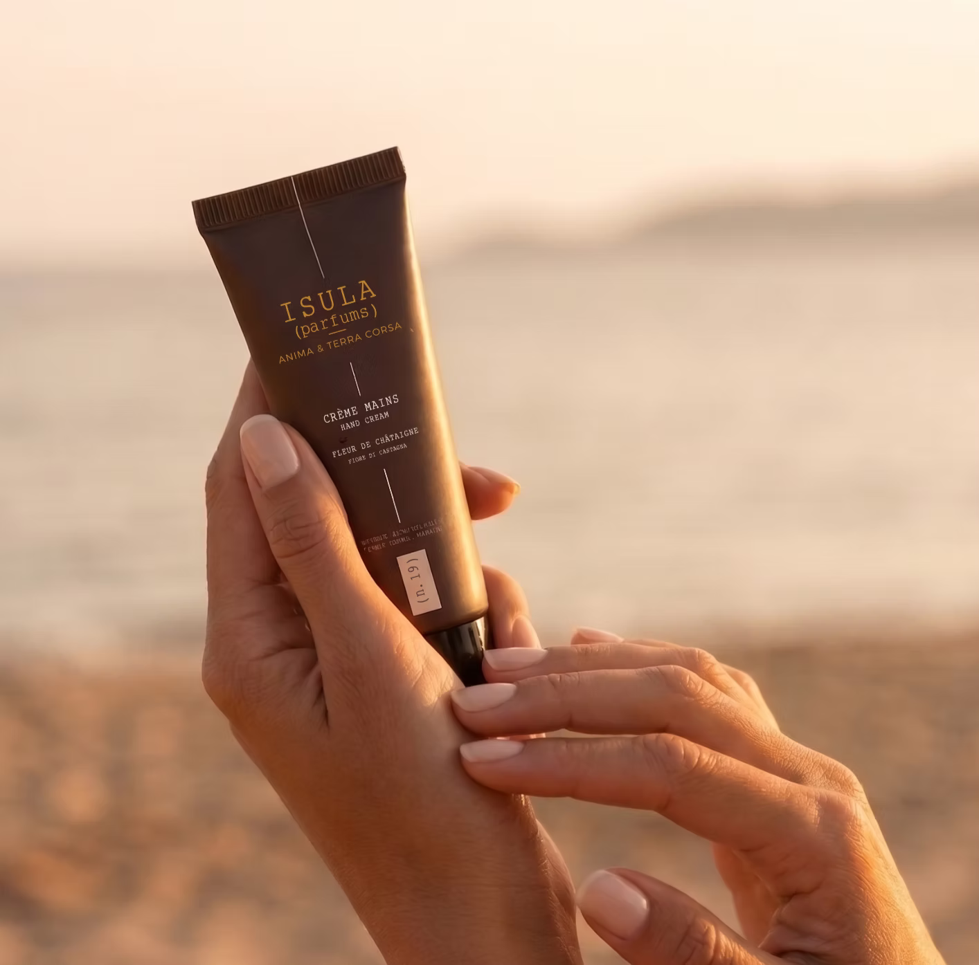 creme à la chataigne qui sent la creme solaire. petit format pour le glisser dans le sac. deux mains de femme qui tiennent le tube de crème, avec un arrière plan flou de plage.