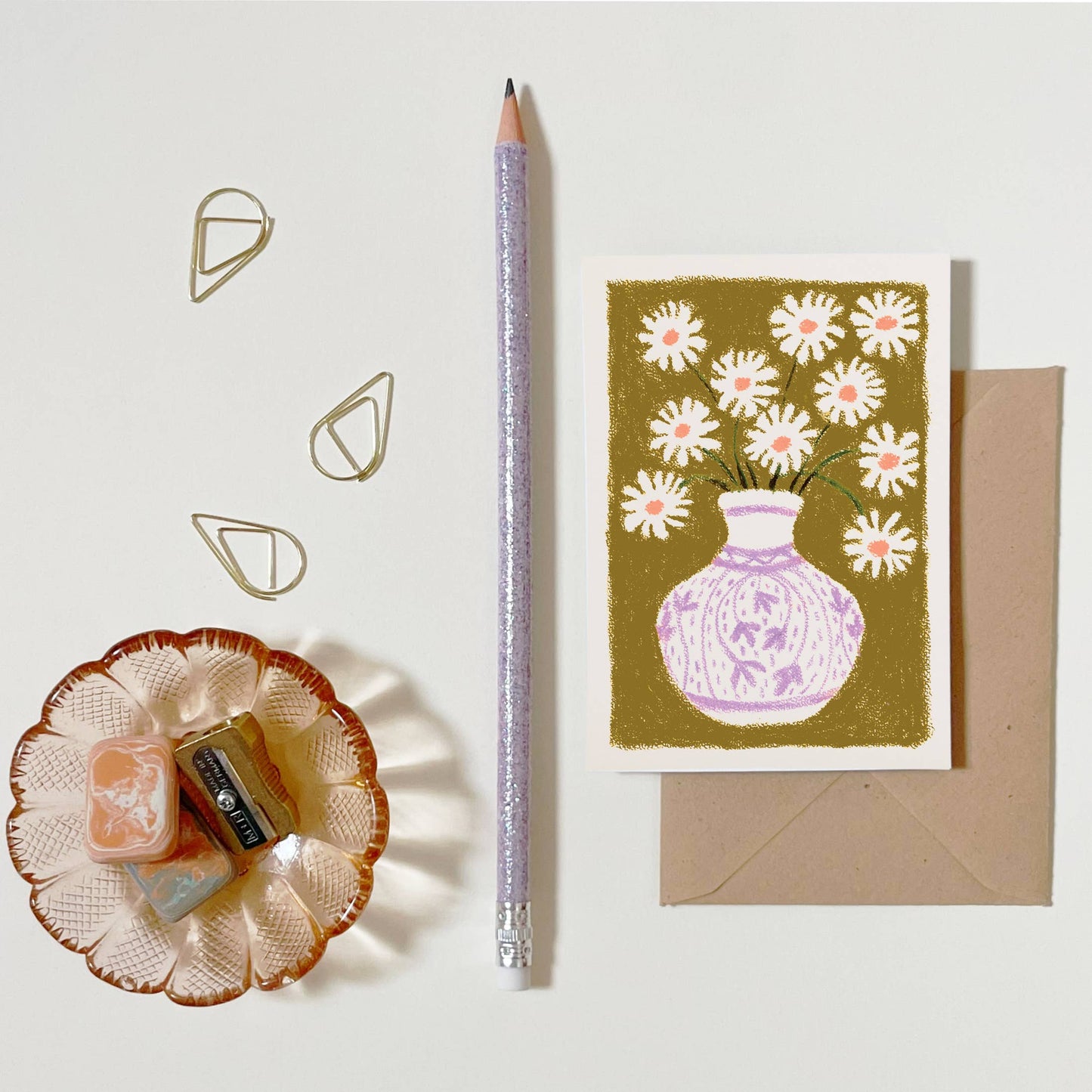 mini carte de voeux avec des marguerites dans un vase, posé sur un fond blanc avec un crayon, un taille crayon et une coupelle