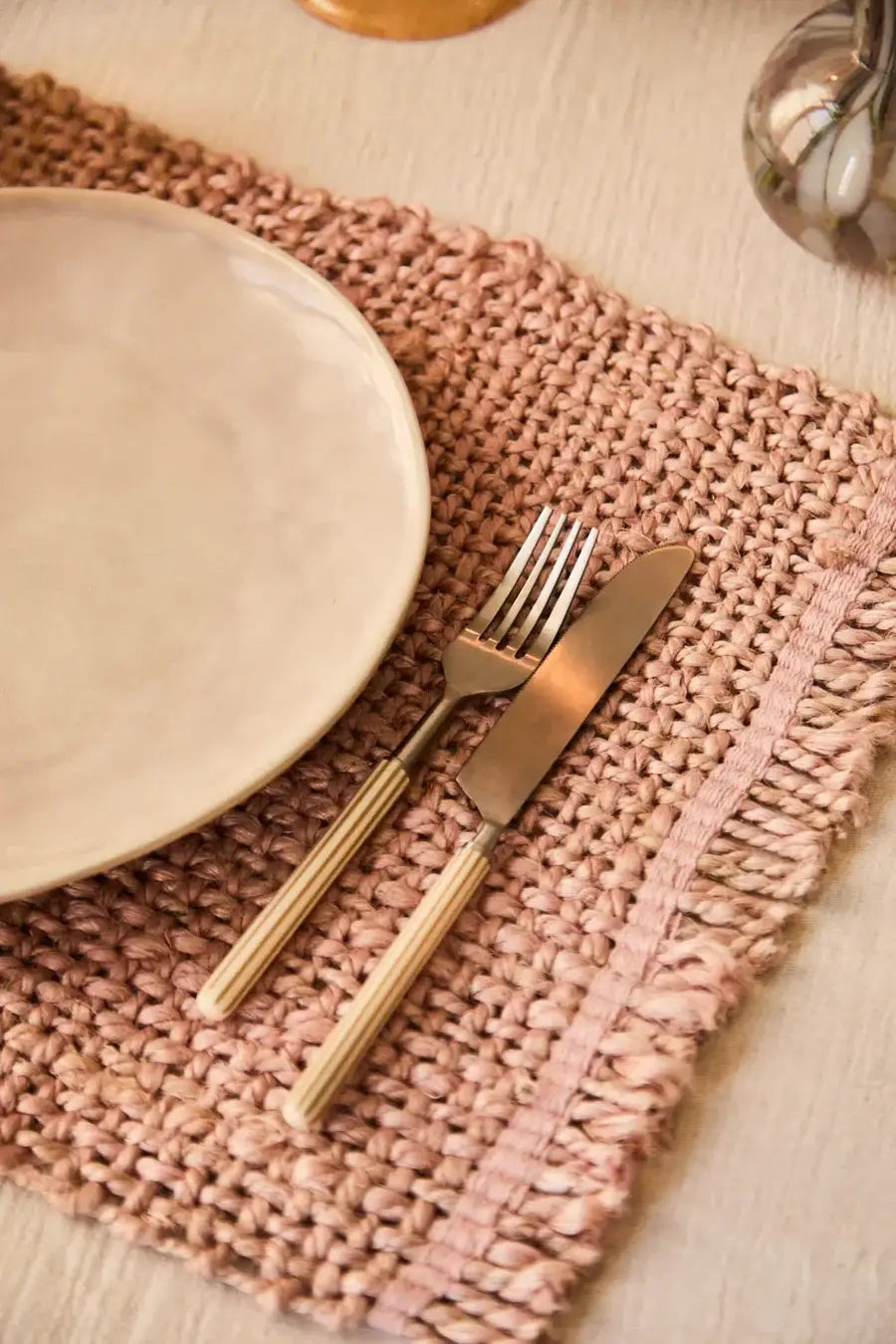 set de table en jute rose avec une assiette beige posée dessus et des couverts avec des rayures