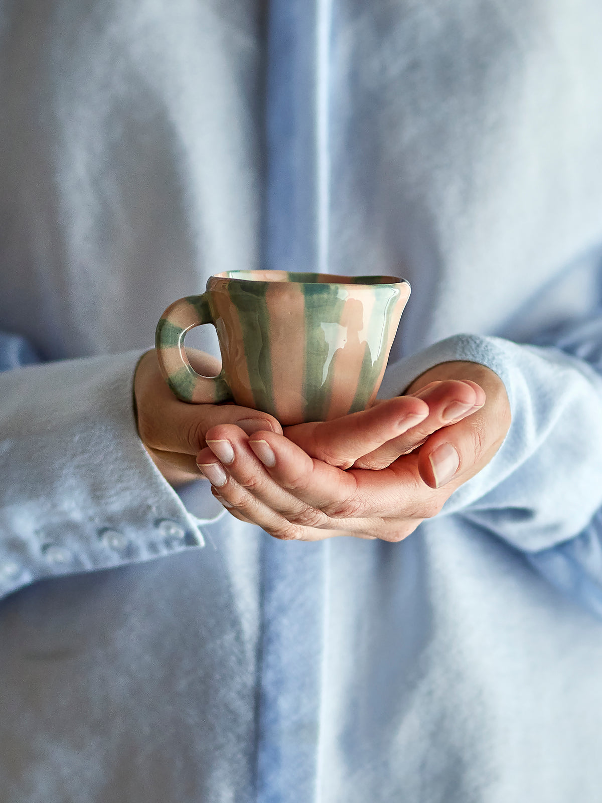 Petite tasse Nini en grès rose peinte à la main avec rayures verticales vert pâle et anse arrondie, contenance 110 ml dans les mains d'une femme